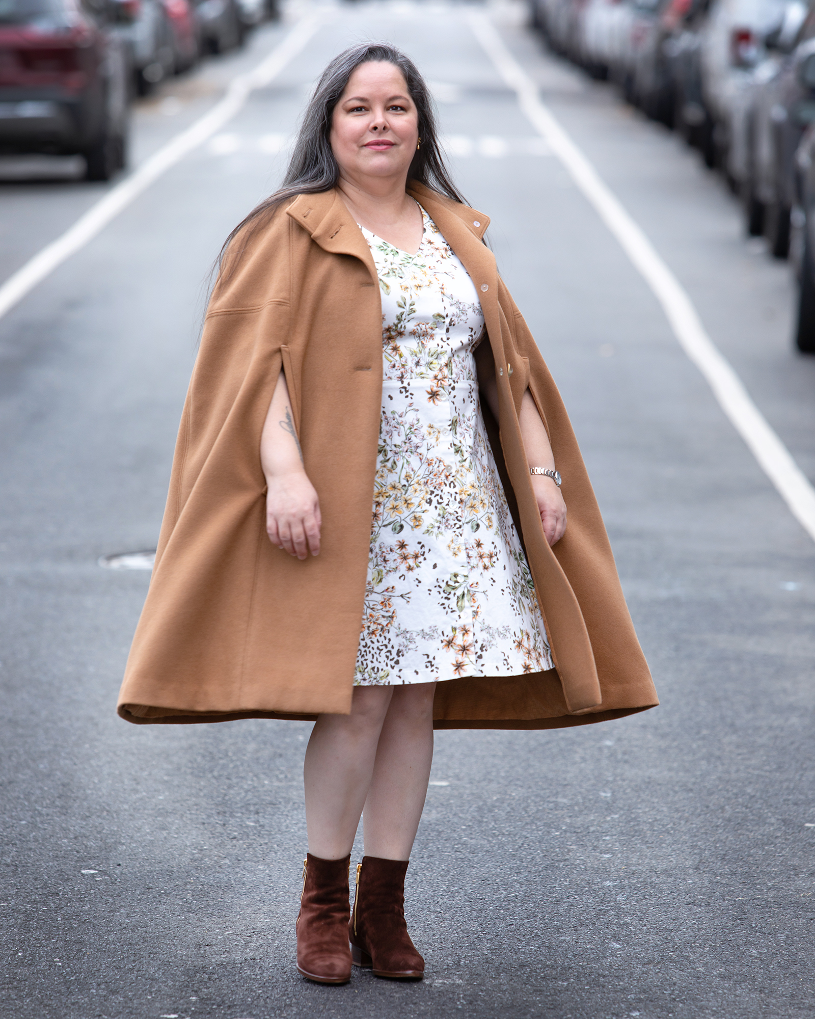 A woman standing in the middle of the street wearing a Burda Style cape over a floral dress with a cream background. She is also wearing brown booties.