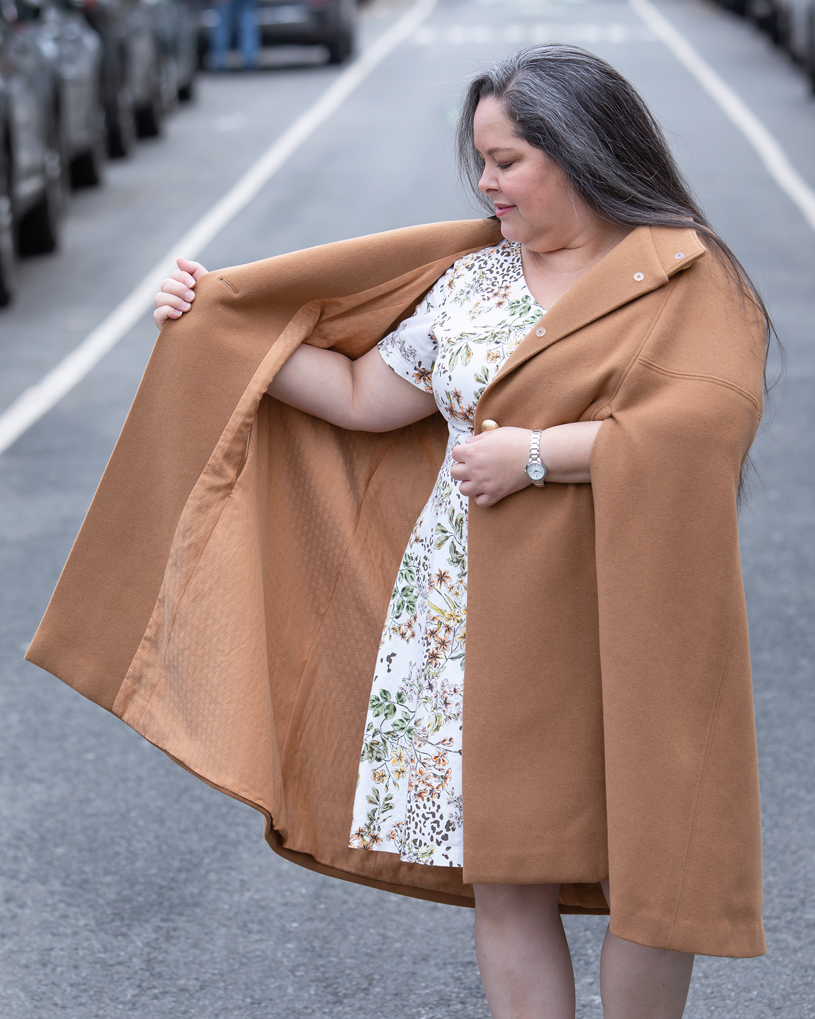 A woman standing in the middle of the street wearing a Burda Style cape over a floral dress with a cream background. She is also wearing brown booties. She is holding the right side of the cape open to show the lining.